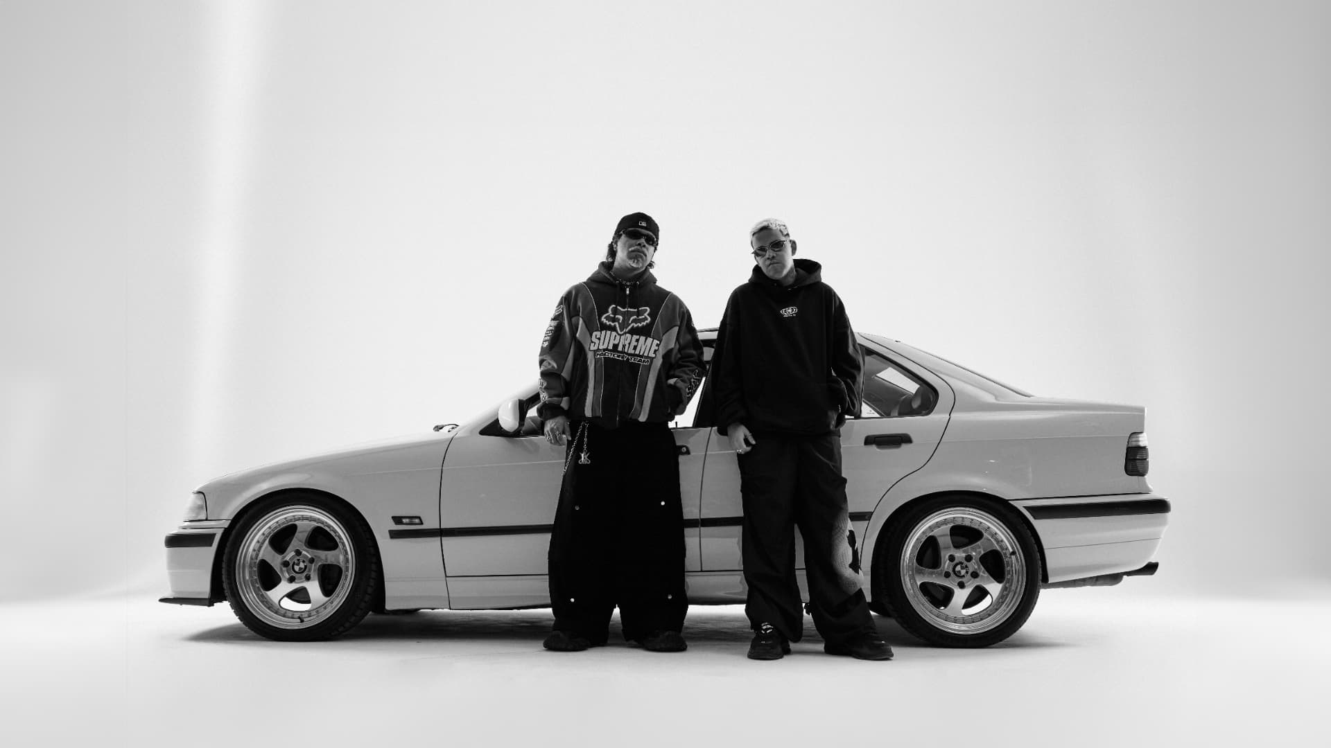 Dois artistas em pé ao lado de um carro antigo no fundo infinito branco do estúdio